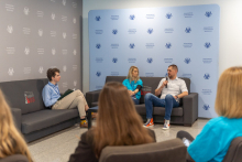 Na planie głównym trzy osoby, siedzą na kanapach i rozmawiają. Za nimi niebieska ścianka z logo WUM.